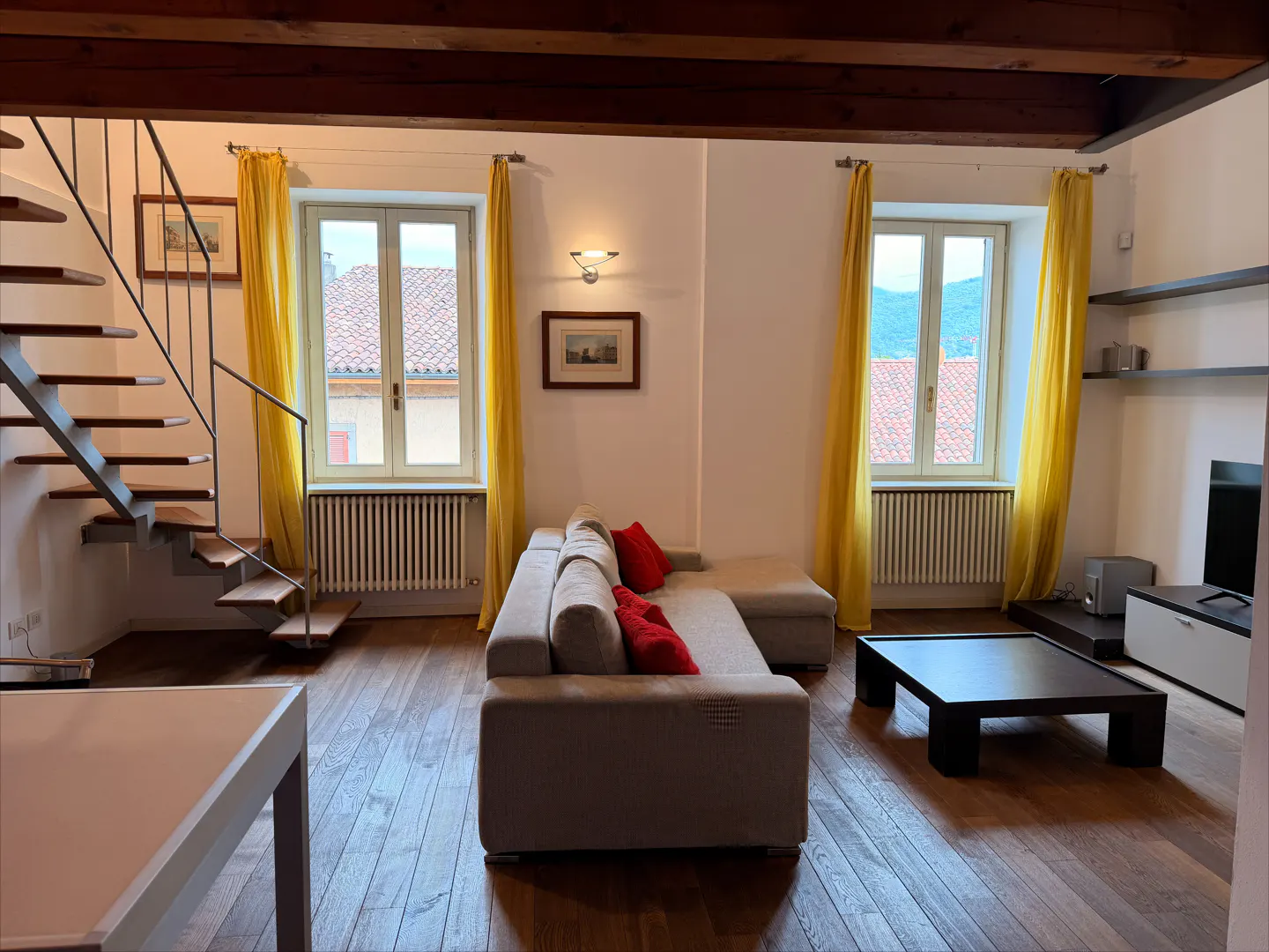 Living room with hardwood floors, beige sectional sofa with red pillows, black coffee table, and metal staircase. Yellow curtains frame two windows.
