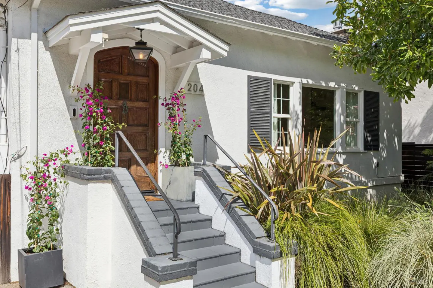 Exterior of a white house with a brown door, gray stairs, and pink flowers.