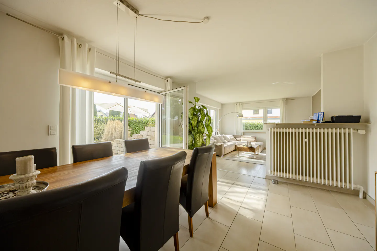 Bright, open-plan dining and living room with a wooden table, leather chairs, and a view to a garden through sliding glass doors.