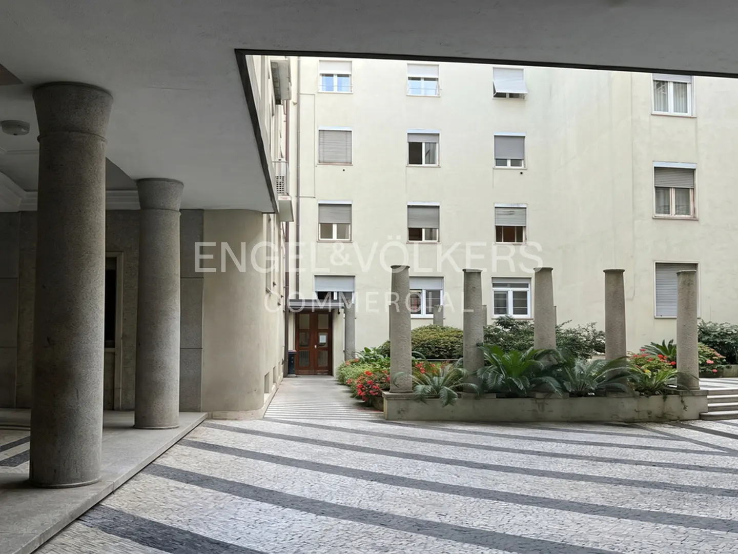 Courtyard view with gray columns, striped pavement, and a building facade with windows and a wooden door.