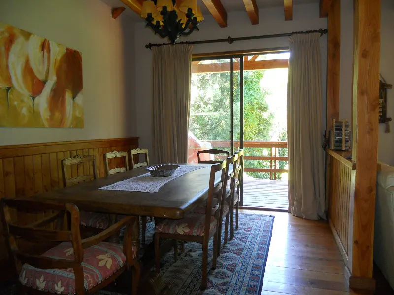 Dining room with wood table, chairs, and rug. Sliding glass doors lead to a deck with green trees in the background.