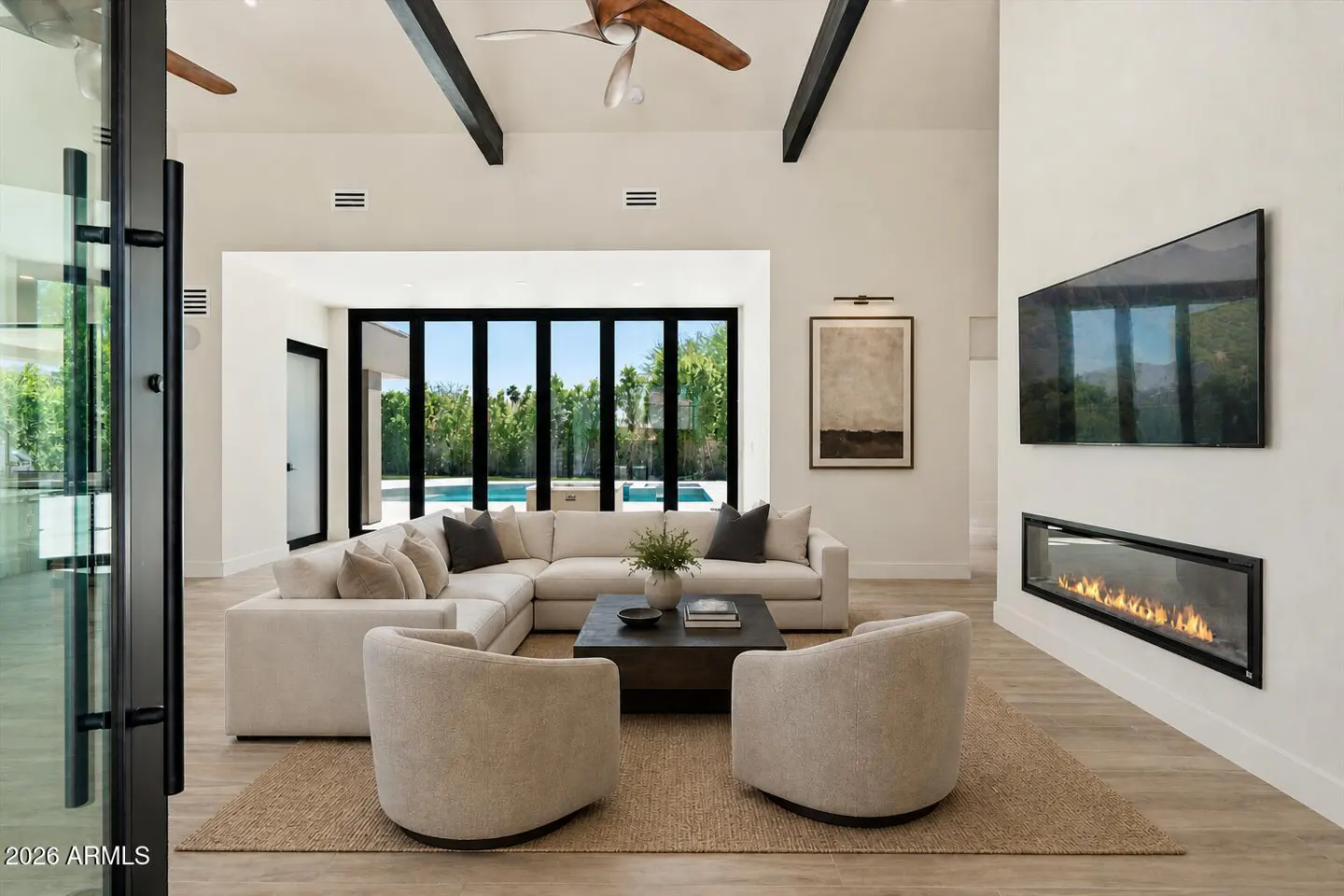 Bright living room with white walls, a fireplace, and a large sectional sofa. Black framed windows overlook a pool.