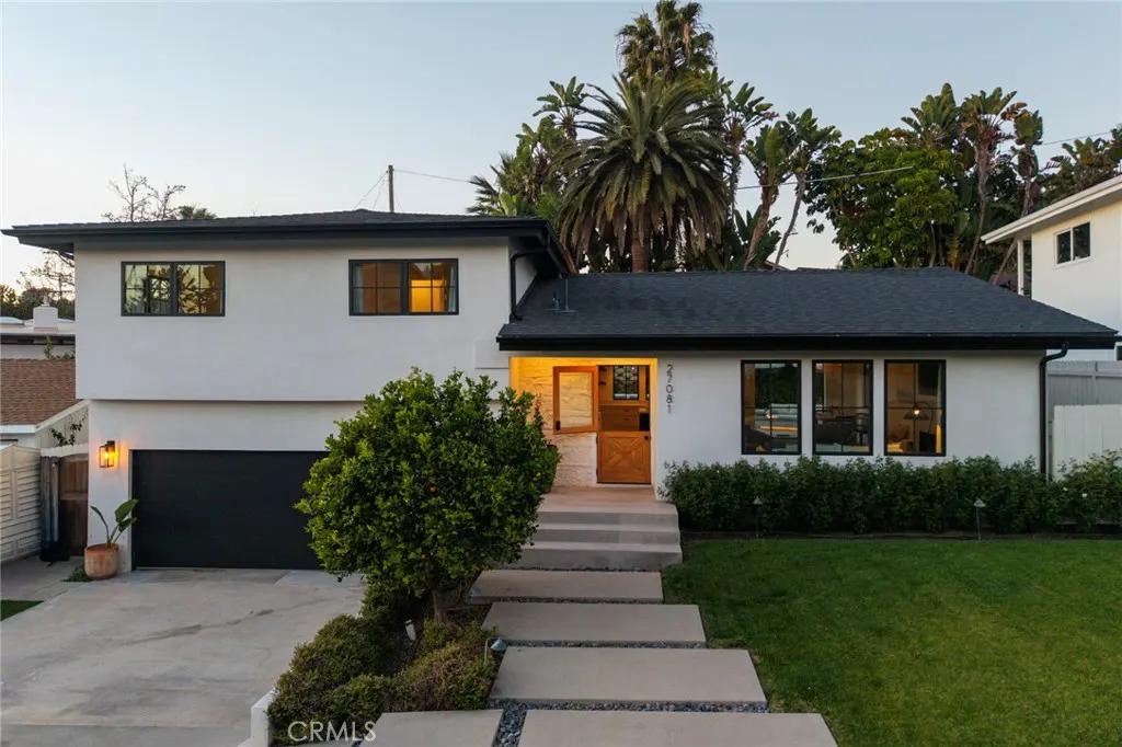 Two-story white house with a black roof and black framed windows. Concrete steps lead to a wooden door. Green lawn and palm trees.