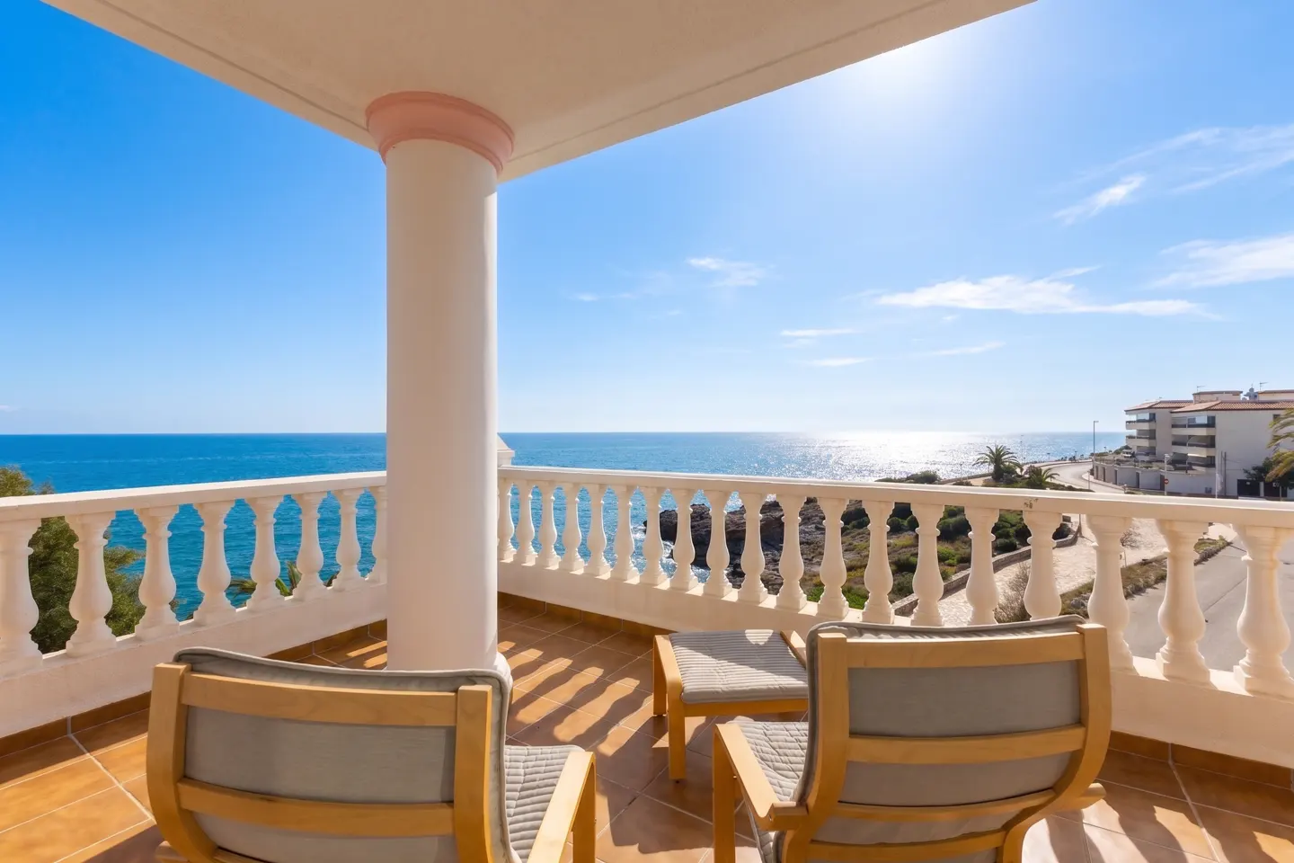 Balcony with two chairs overlooking the ocean. White pillars and railing frame the blue sea and sky.