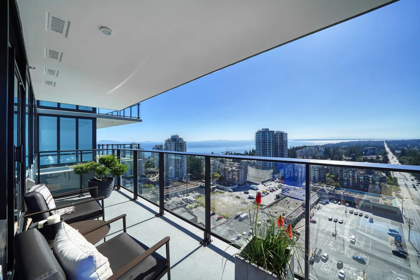 Condo balcony with glass railings, chairs, and potted plants overlooking a city skyline and ocean on a sunny day.