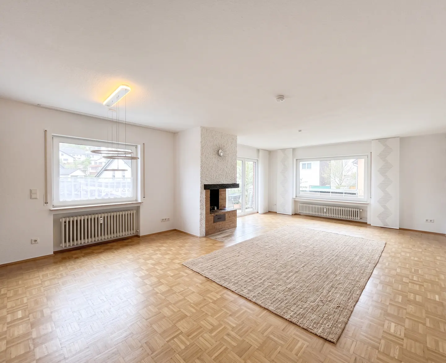 Bright, empty living room with parquet floors, a beige rug, and a fireplace. Windows offer outdoor views.