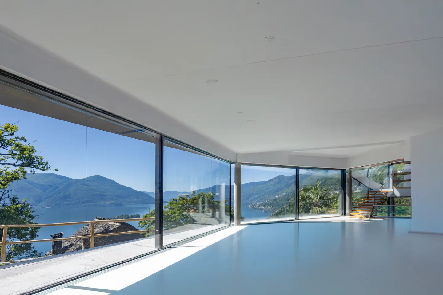 Bright, empty room with white walls and floor. Large windows offer a scenic view of a lake and mountains. Stairs are visible in the background.