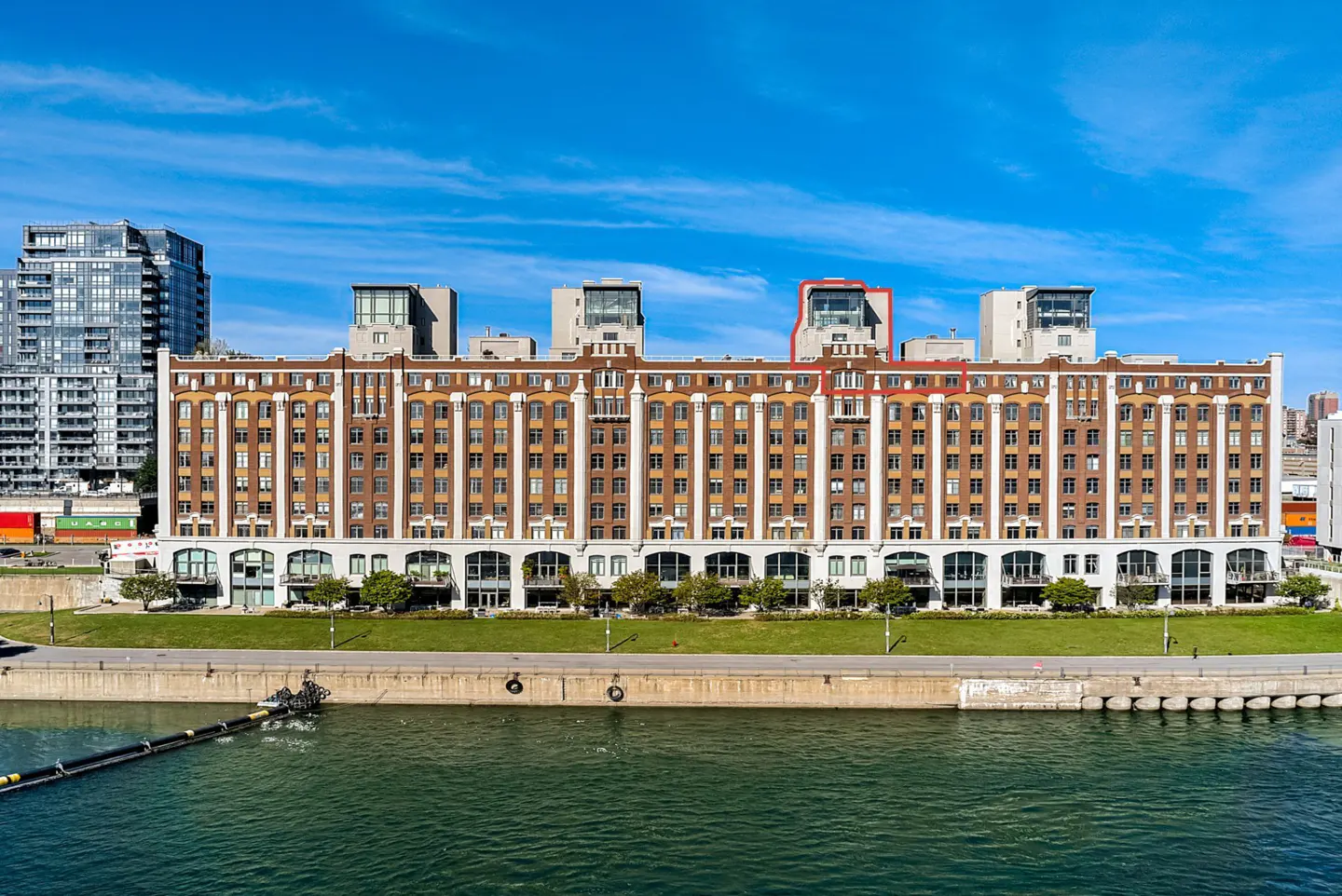 Exterior view of a brown and white brick apartment building with a green lawn, blue sky, and a body of water in front.