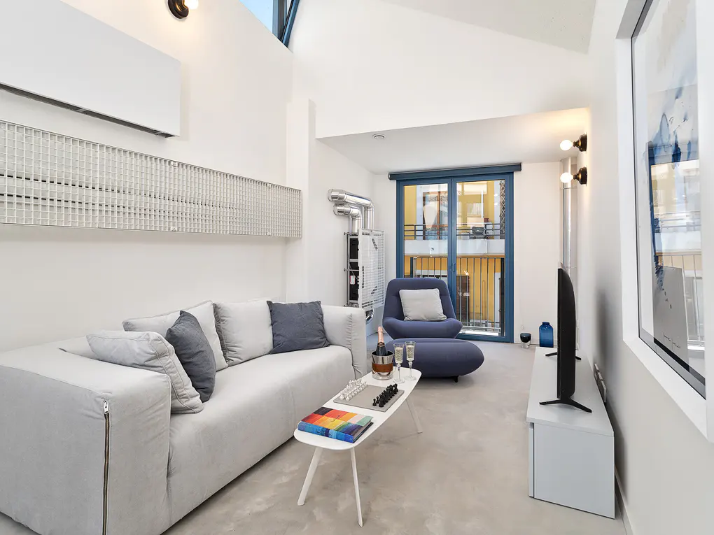 Bright, modern living room with white walls, a gray sofa, and a blue chair. A white coffee table holds books and a chess set. Balcony view.