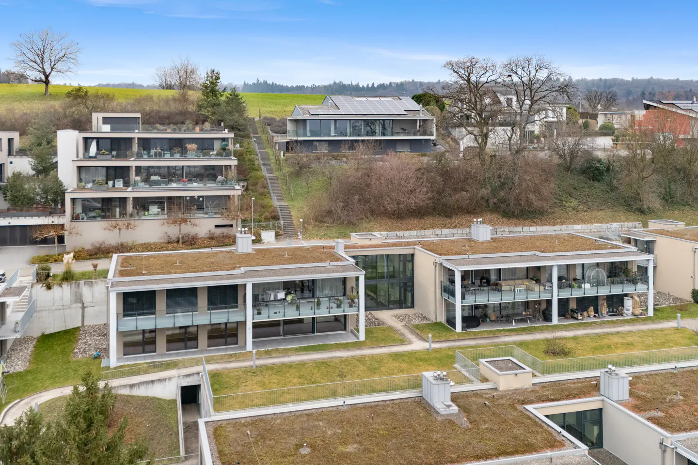 Aerial view of modern apartments with green roofs and balconies, set on a grassy hillside under a blue sky.