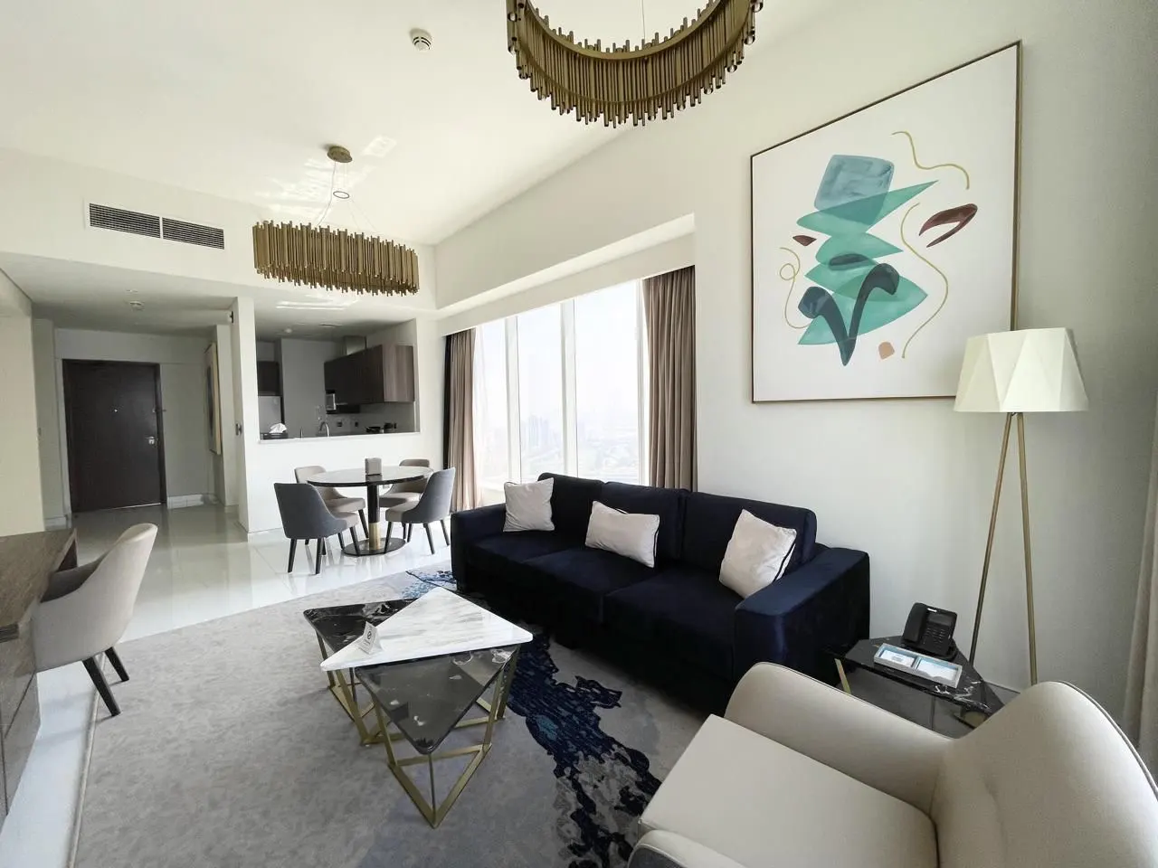 Bright, modern apartment living room with blue sofa, white armchair, and abstract art. Dining area and kitchen visible in the background.