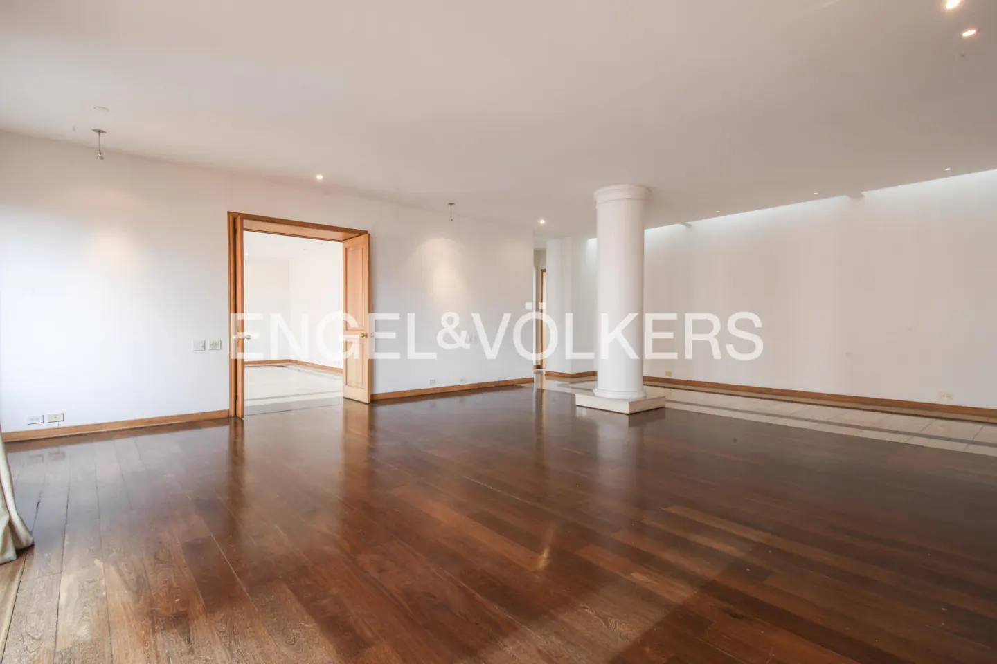 An empty room with hardwood floors, white walls, and a white pillar. A doorway is visible in the background.