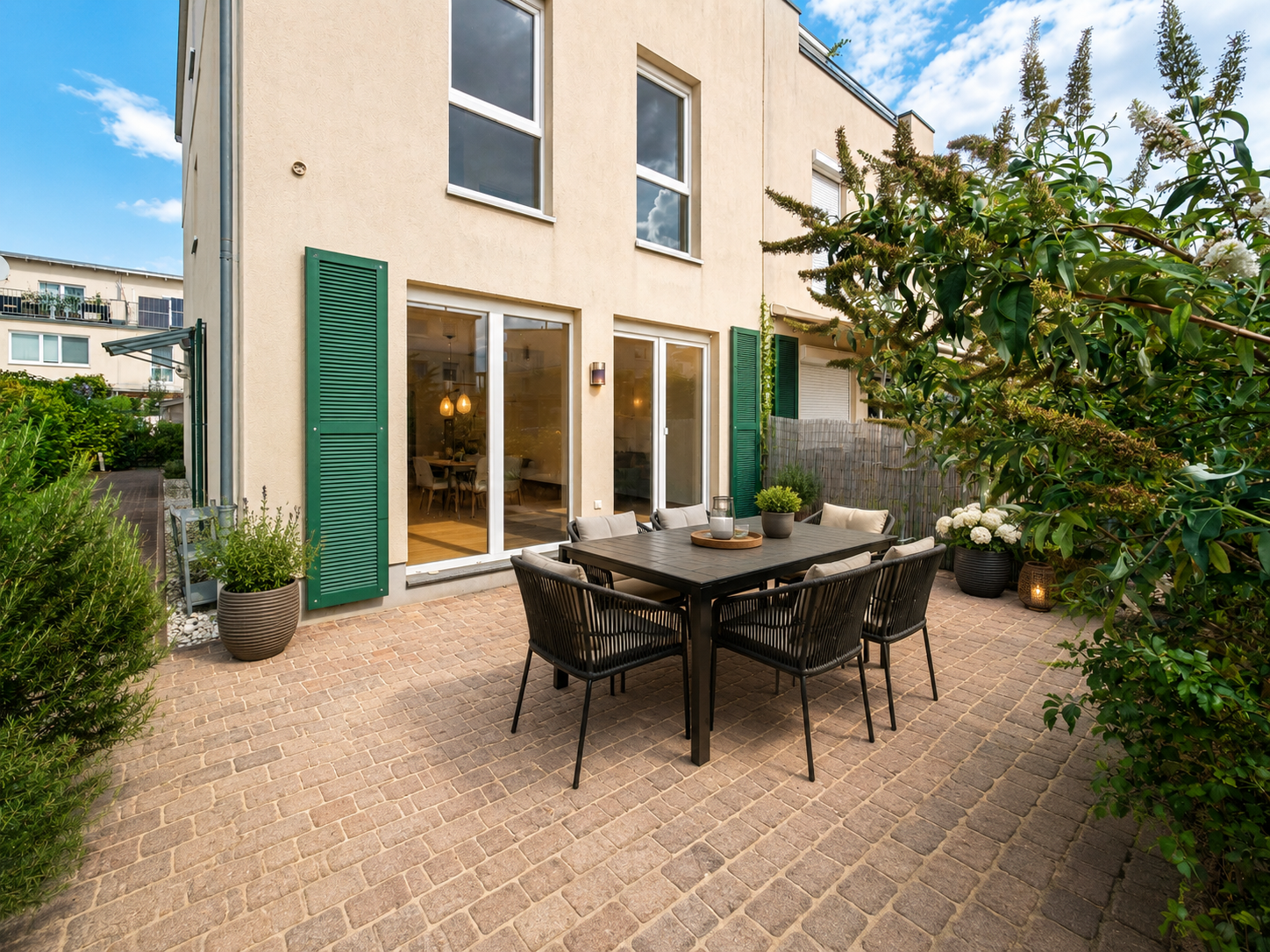 A beige house exterior with a paved patio, outdoor dining set, green shutters, and lush garden under a blue sky. [AI-generated text]
