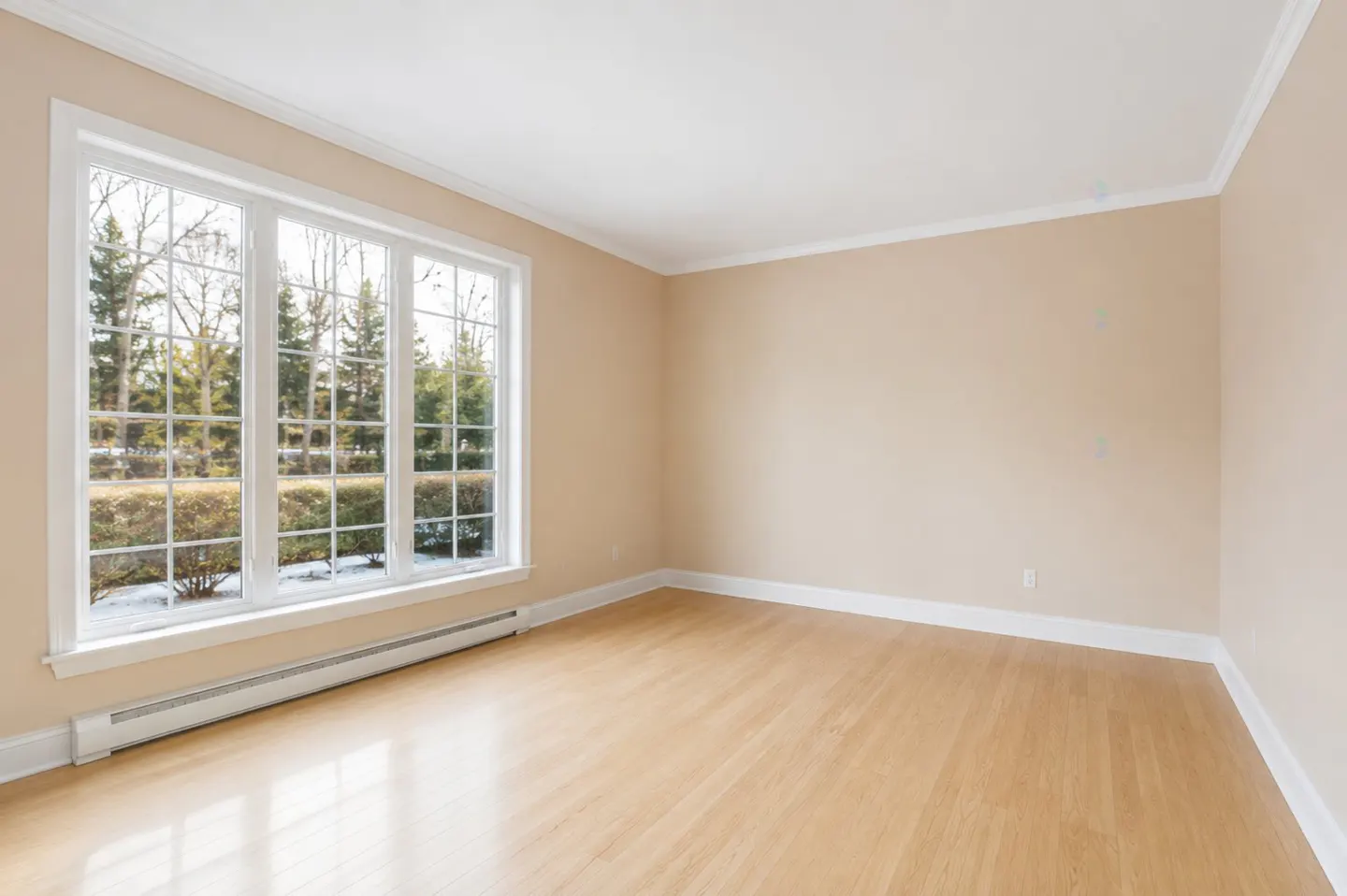 Empty room with light wood floors, beige walls, and a large window with a view of trees and snow.
