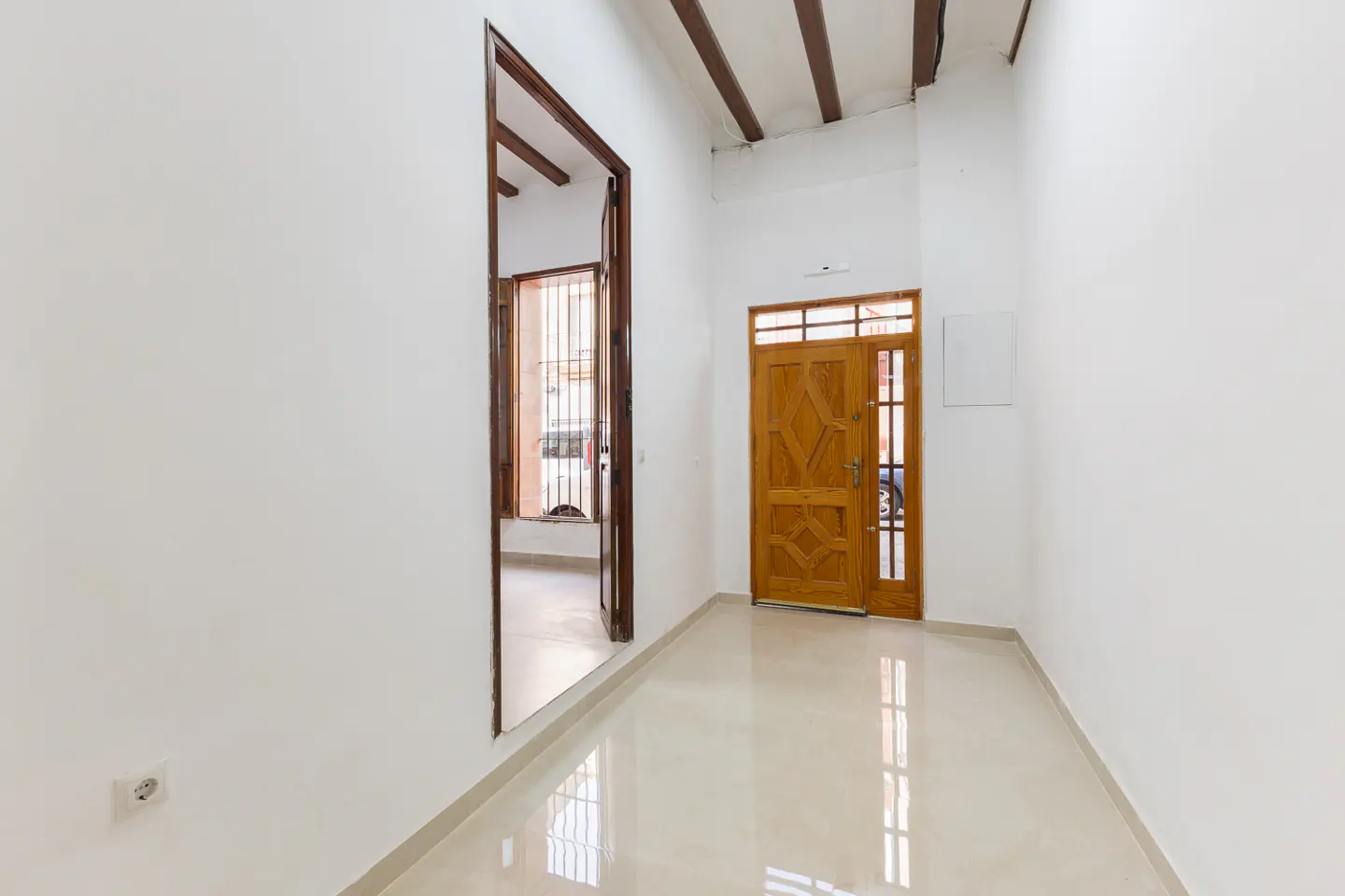 Bright hallway with white walls, beige tile floor, and wooden doors. A doorway leads to another room with a window. Ceiling has exposed wooden beams.
