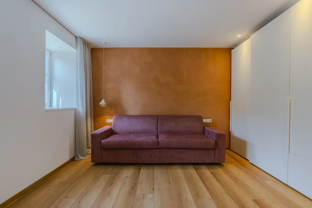 A modern room with a maroon sofa against an orange wall, light wood floors, and white closet doors. A window with sheer curtains is on the left.
