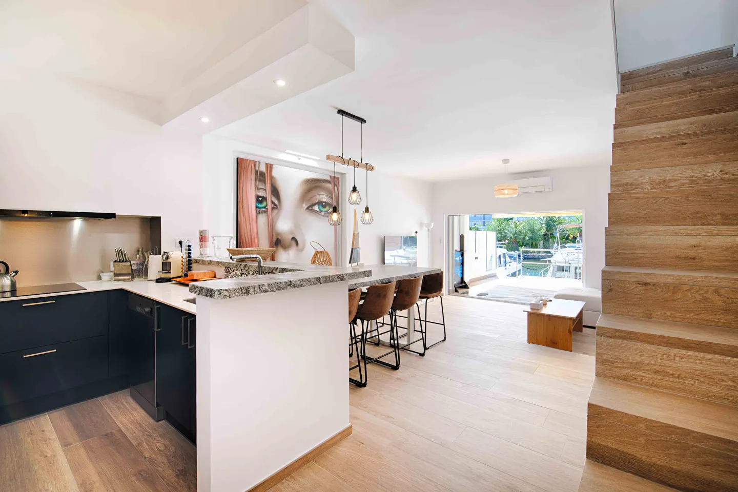Bright, modern kitchen and living area with wood floors, black cabinets, and a large painting of a woman's face. Stairs lead to the upper level.