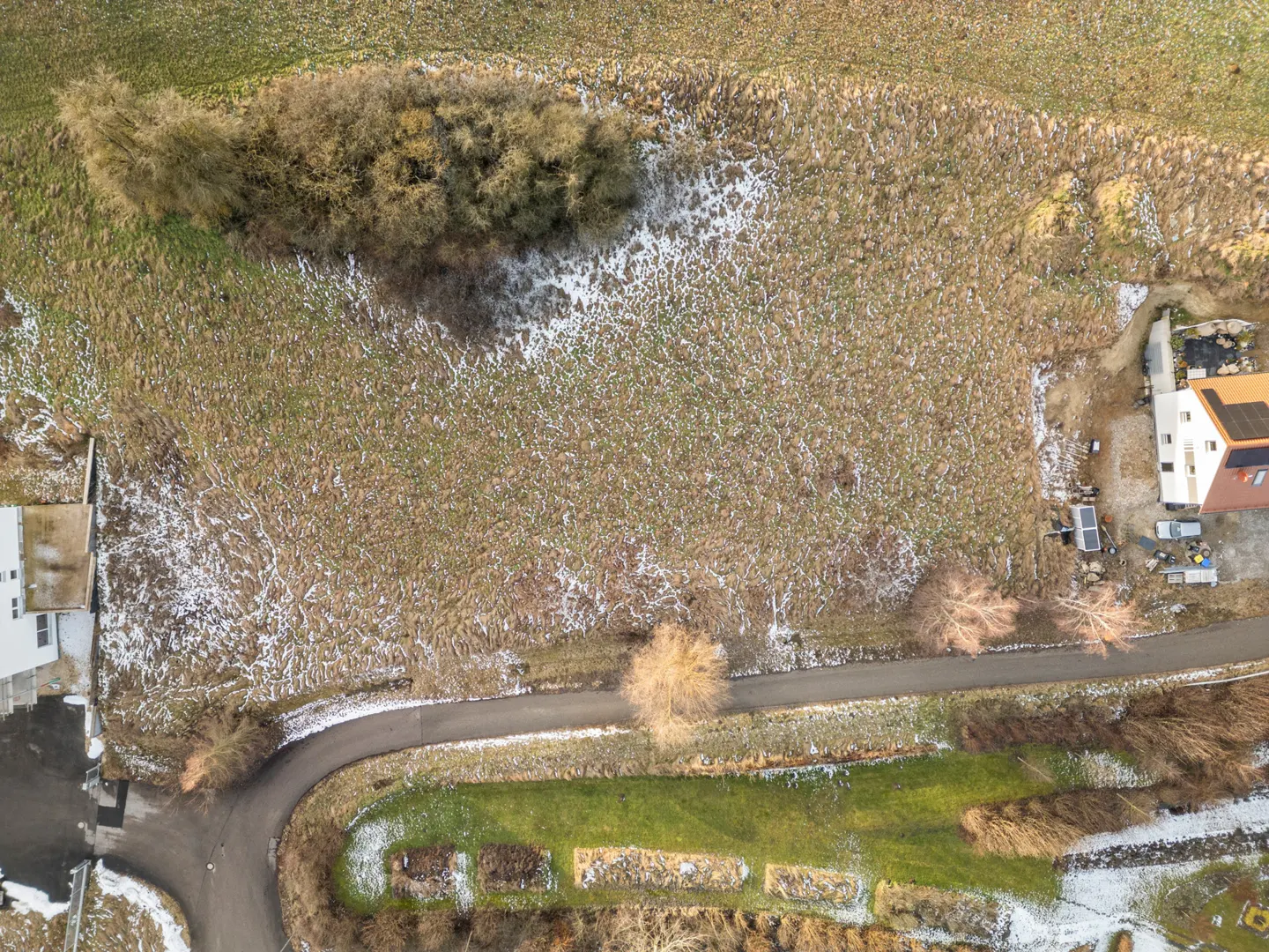 Aerial view of a vacant lot with sparse snow cover, bordered by trees, a road, and houses.