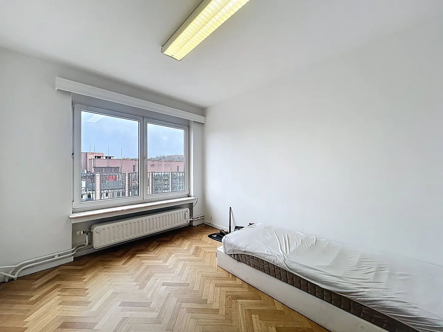 Bright, empty room with herringbone wood floors, white walls, and a large window overlooking city buildings. A bed with a plastic cover sits in the corner.