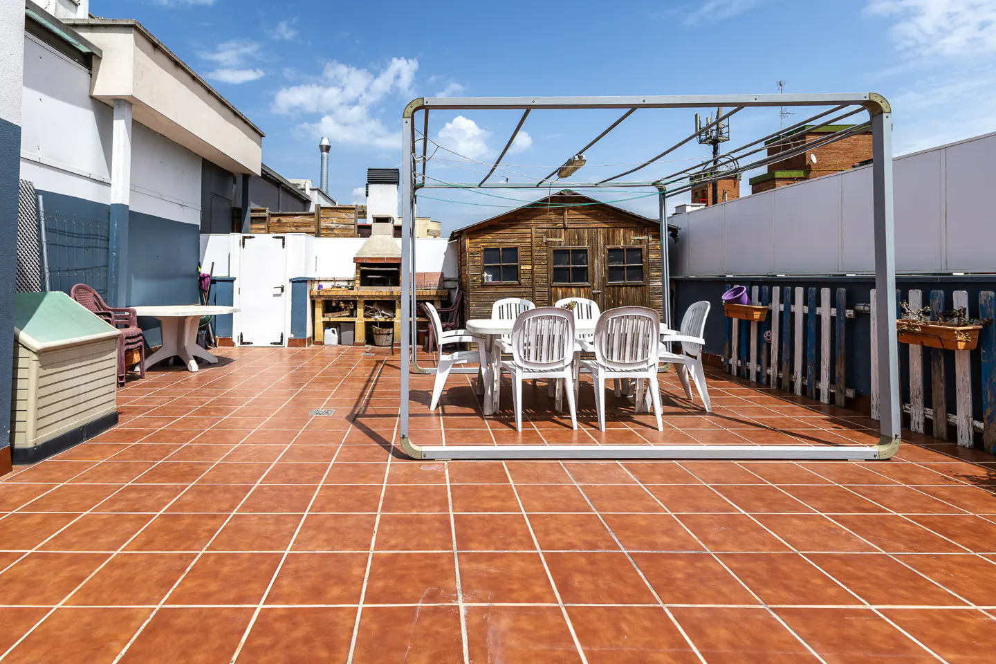 Rooftop patio with brown tile, white table and chairs under a metal frame. A wooden shed and brick grill are in the background.
