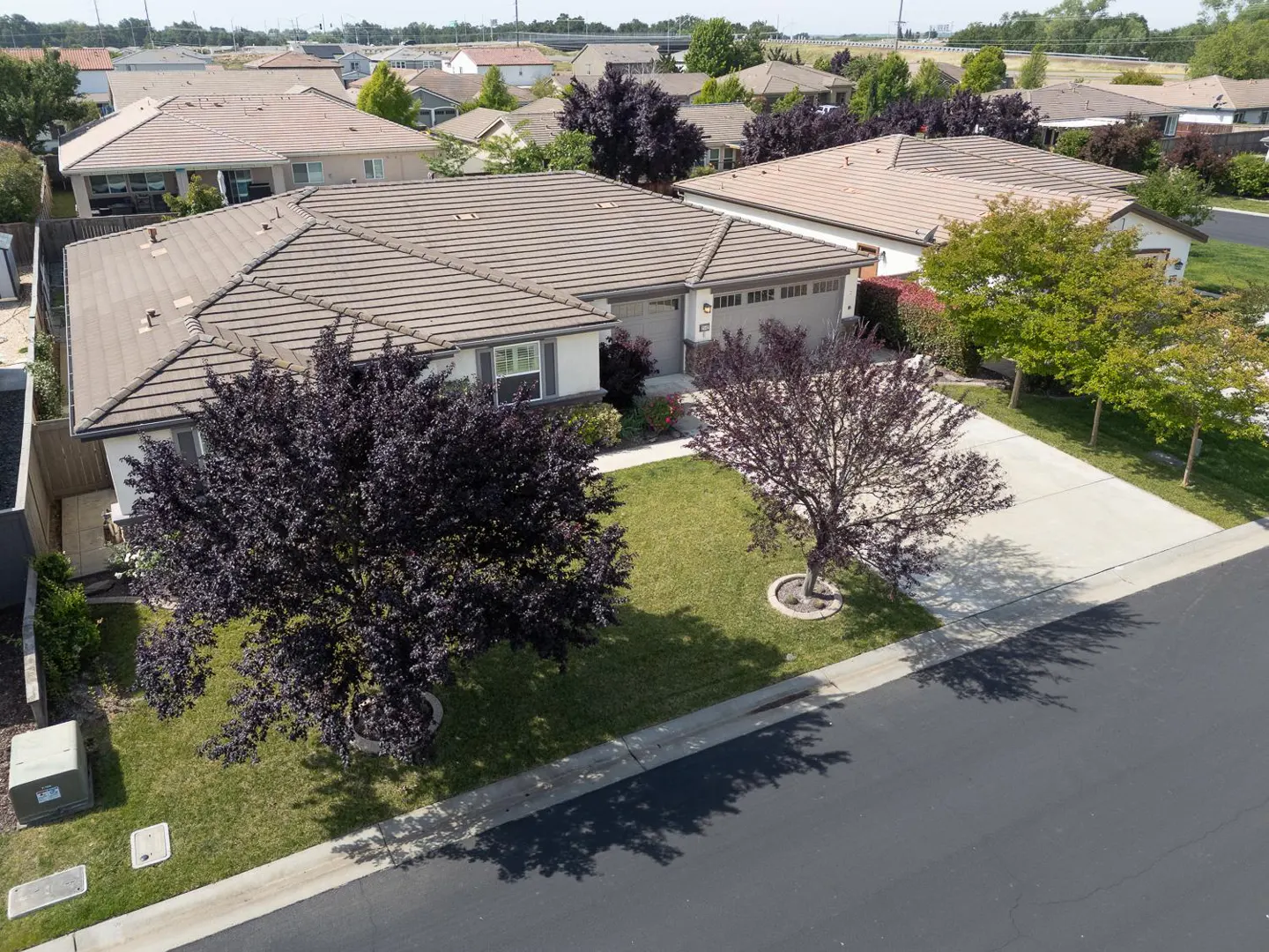 Aerial view of a single-story house with a brown tile roof, gray siding, and a two-car garage in a suburban neighborhood.