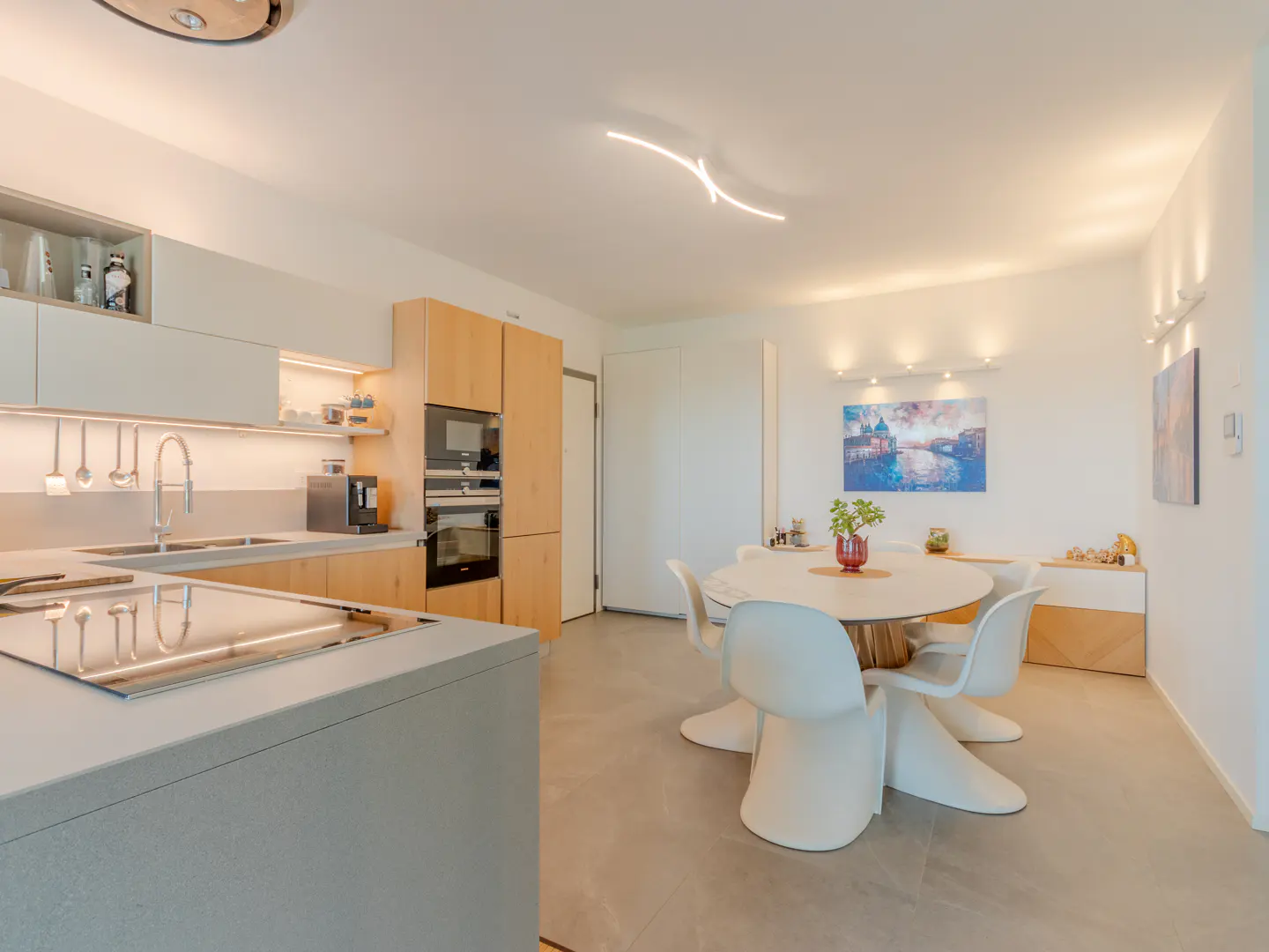 Bright, modern kitchen with light wood cabinets, gray countertops, and a round dining table with white chairs. Artwork adorns the white walls.