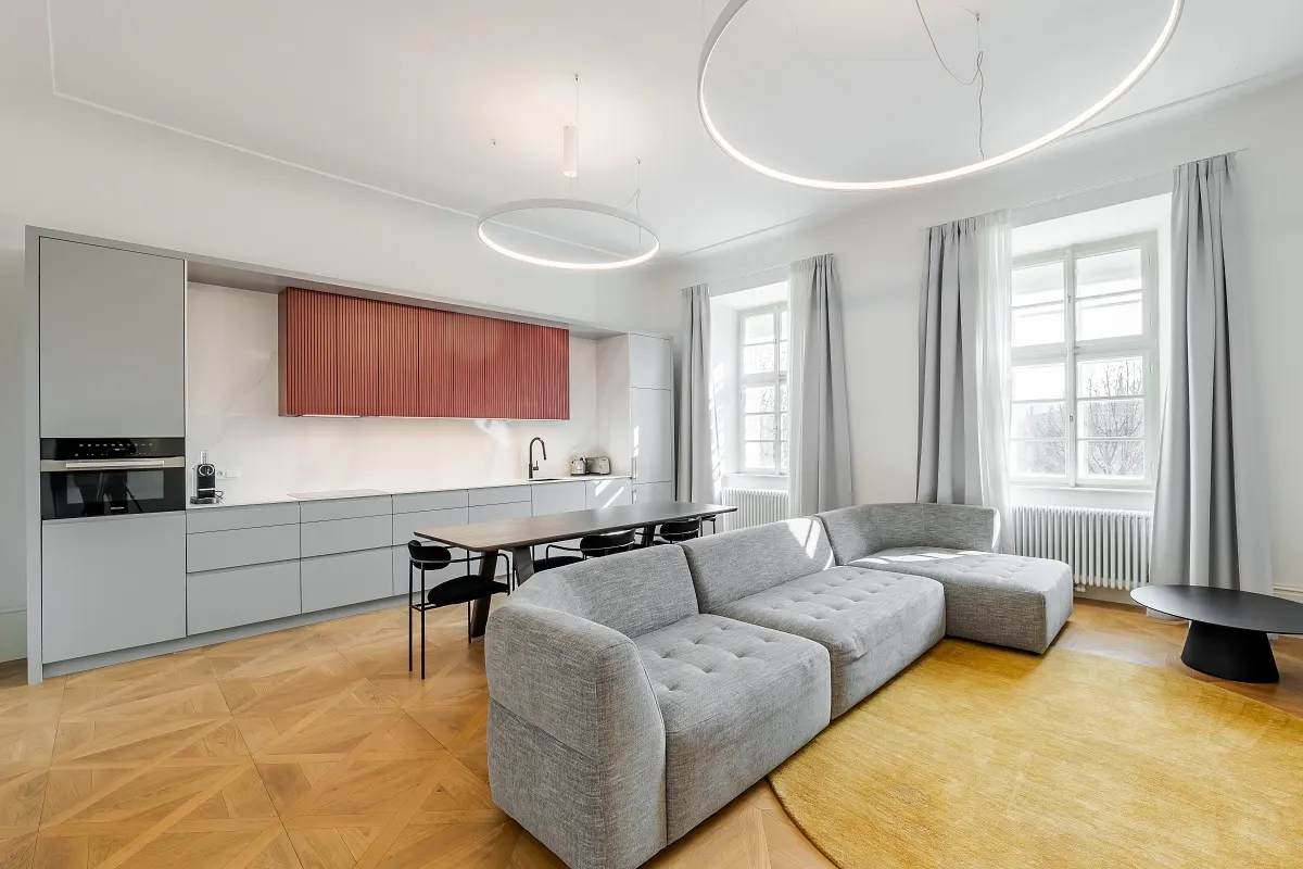 Modern open-plan living area with a grey sofa, yellow rug, dining table, and kitchen with red cabinets and wood floors.