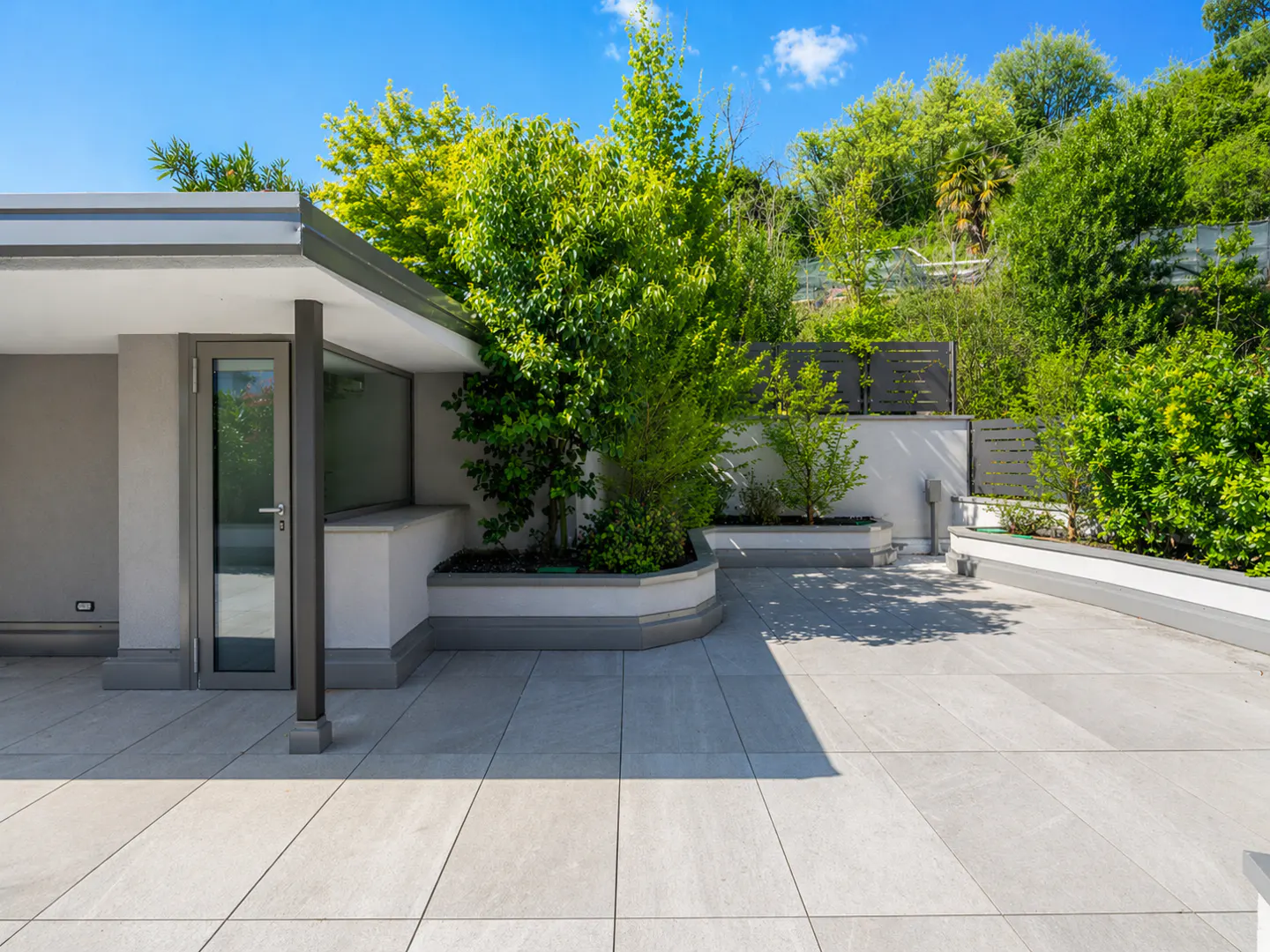 A modern patio with gray tiles, a gray door, and green trees in planters against a lush green hillside.
