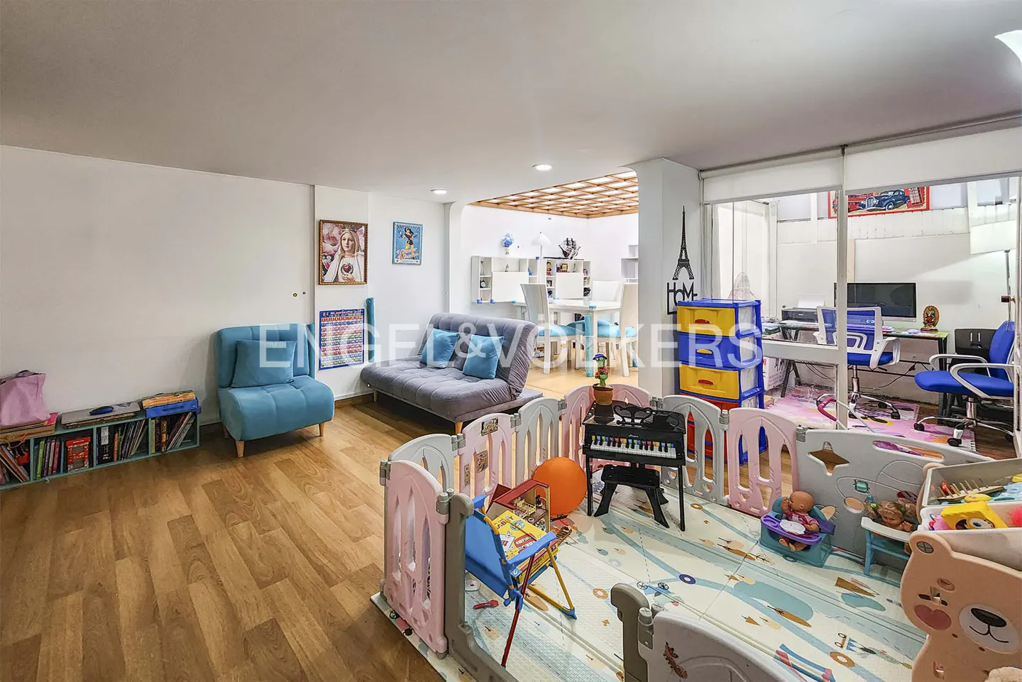 Bright, open playroom with hardwood floors, a blue sofa, and a play area enclosed by a pink and gray fence. Toys and furniture are scattered throughout.