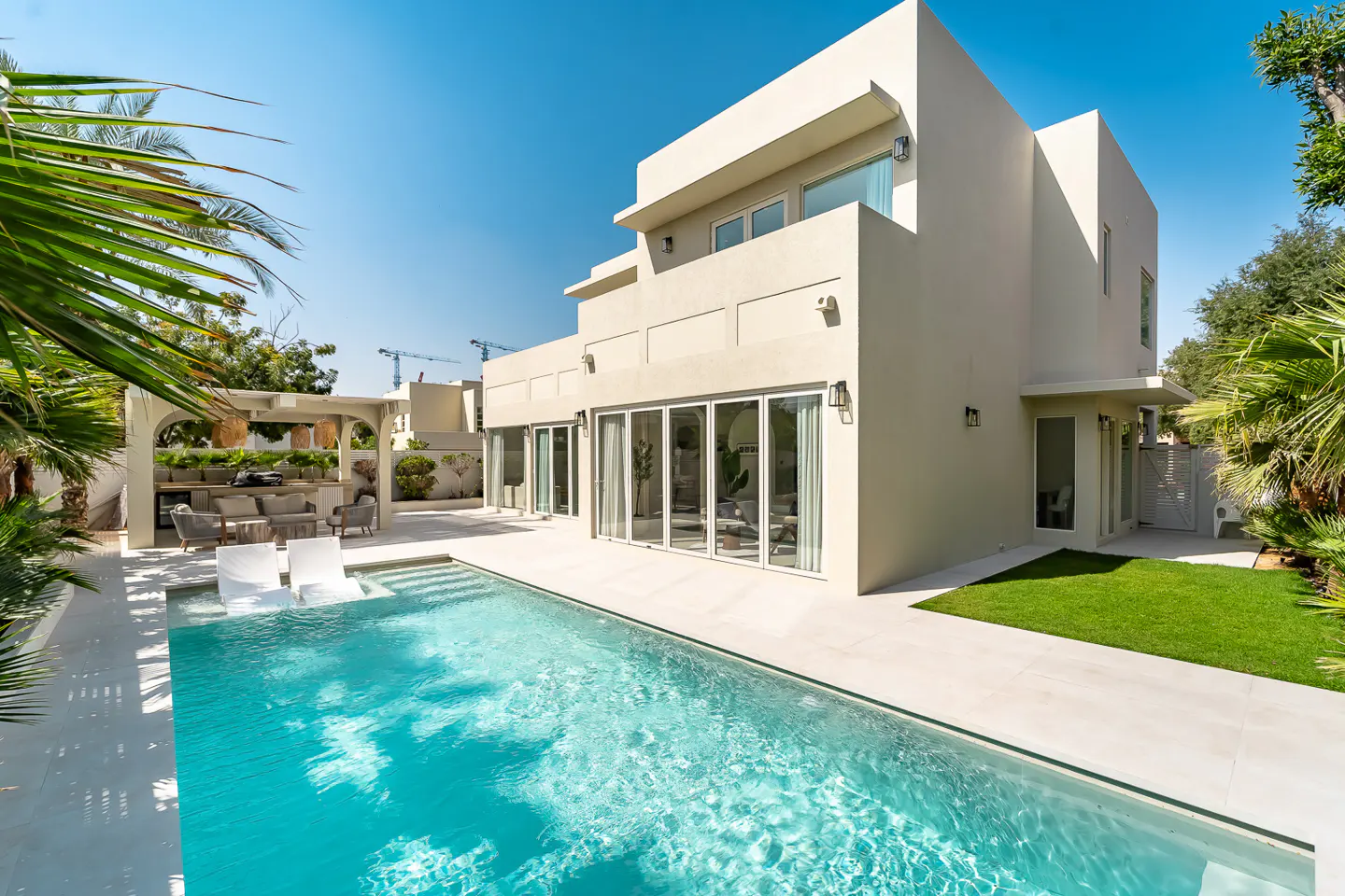 Backyard view of a modern, two-story beige house with a blue swimming pool, outdoor kitchen, and green lawn.