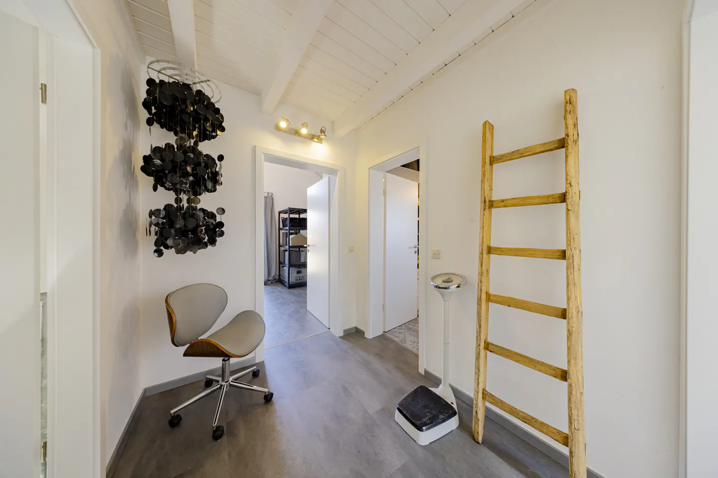 Hallway with gray floors, white walls, and a white ceiling. A gray chair, black chandelier, and wooden ladder decorate the space.