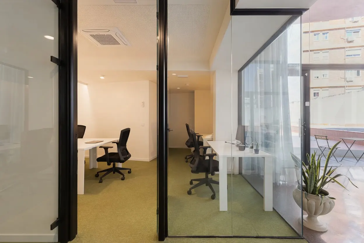 Modern office interior with glass partitions, white desks, black chairs, and green carpeting, overlooking a patio.