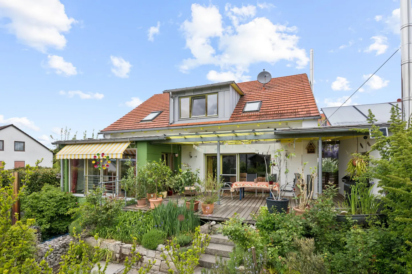 Backyard view of a two-story house with a red-tiled roof, a wooden deck, and lush greenery. The house has a green accent wall and a yellow-striped awning.