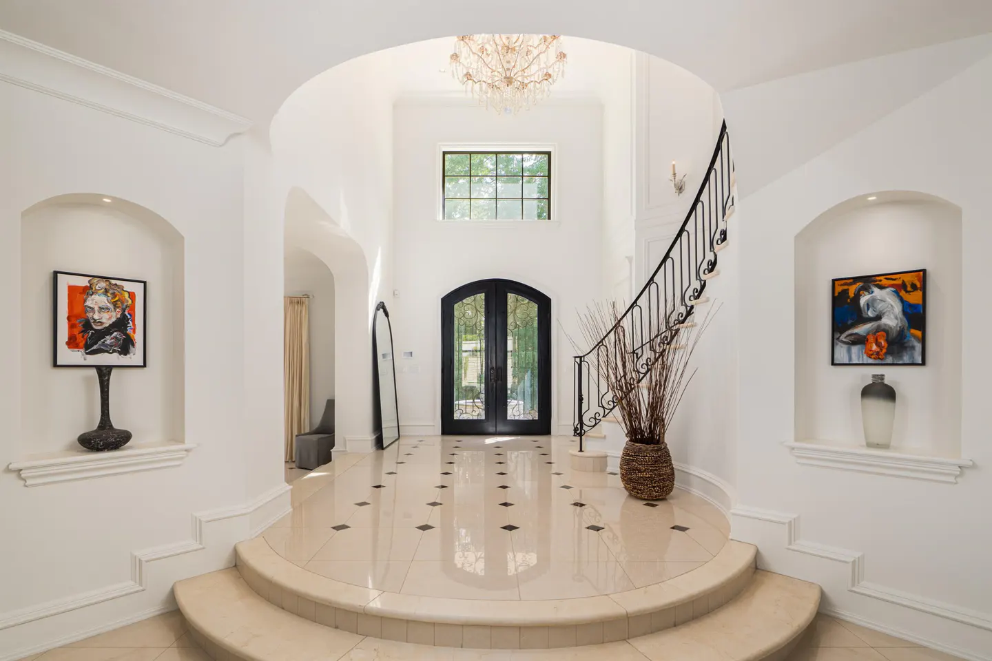 Bright foyer with white walls, beige tile floor, black double doors, and a curved staircase with black iron railing. Artwork in wall niches.