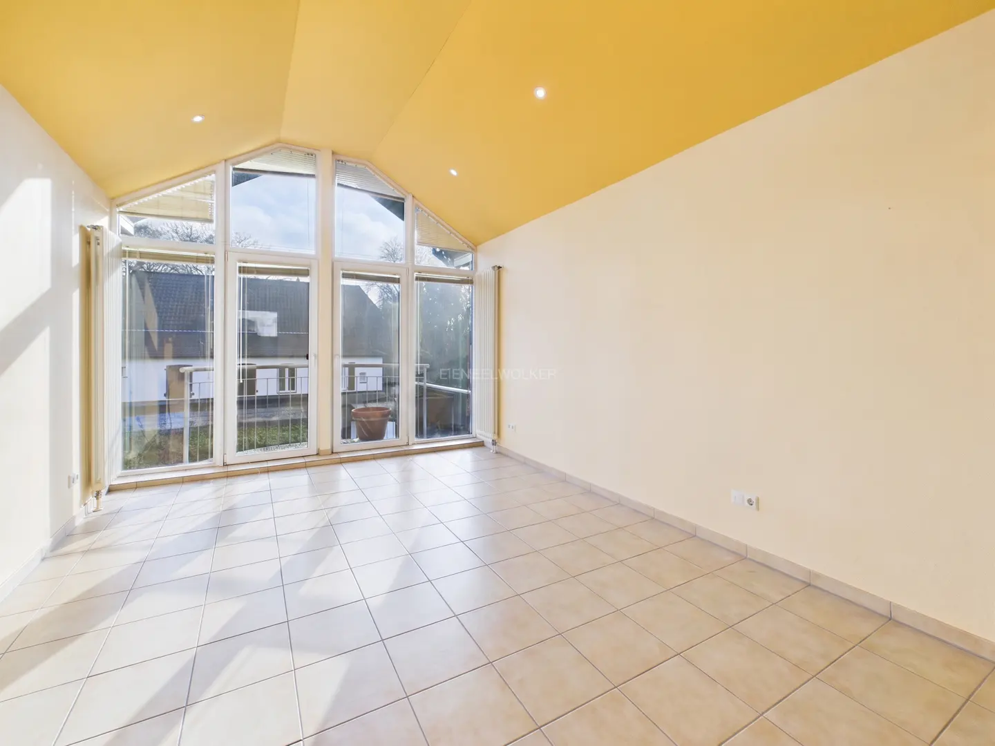 Bright, empty room with yellow ceiling, cream walls, and tiled floor. Large windows offer a view of a house and greenery.