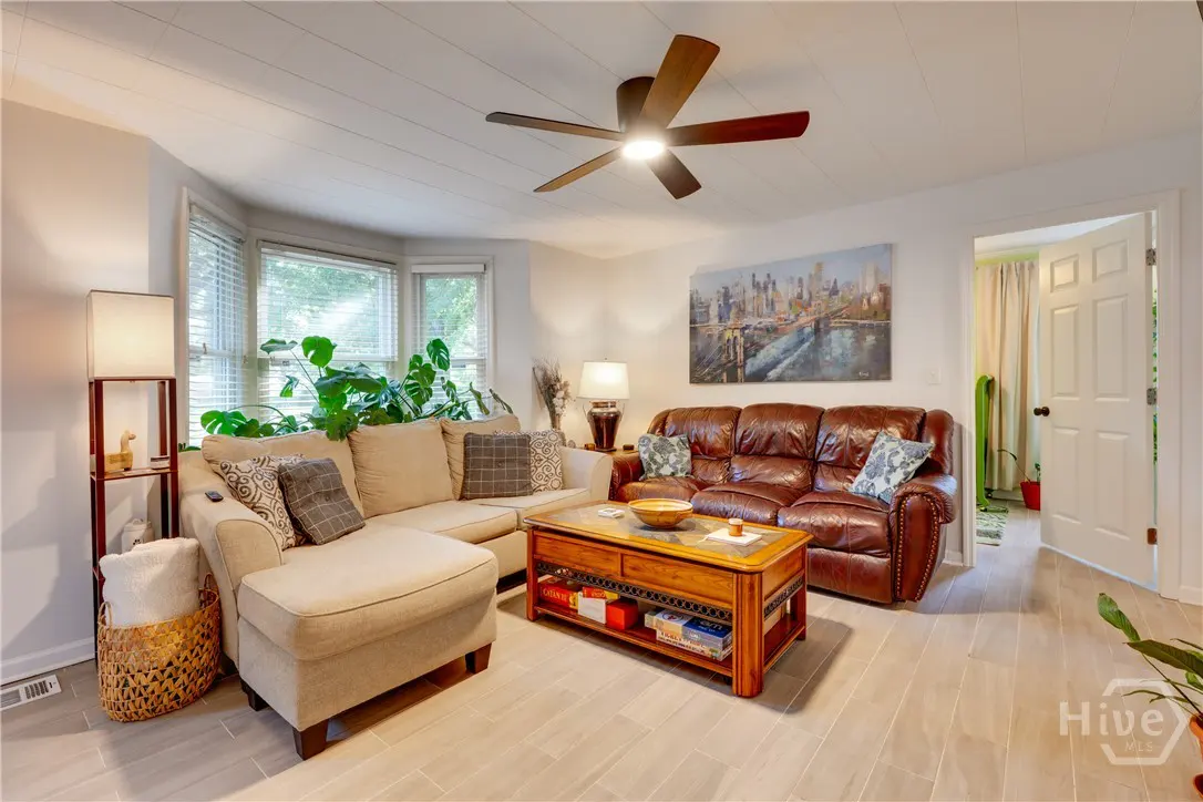 Living room with beige sectional, brown leather sofa, wooden coffee table, ceiling fan, and city skyline wall art.