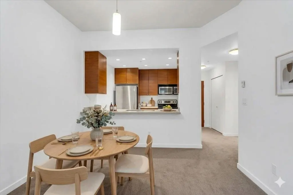 Bright apartment dining area with a round wood table set for four, a vase of flowers, and a view into the kitchen with wood cabinets.
