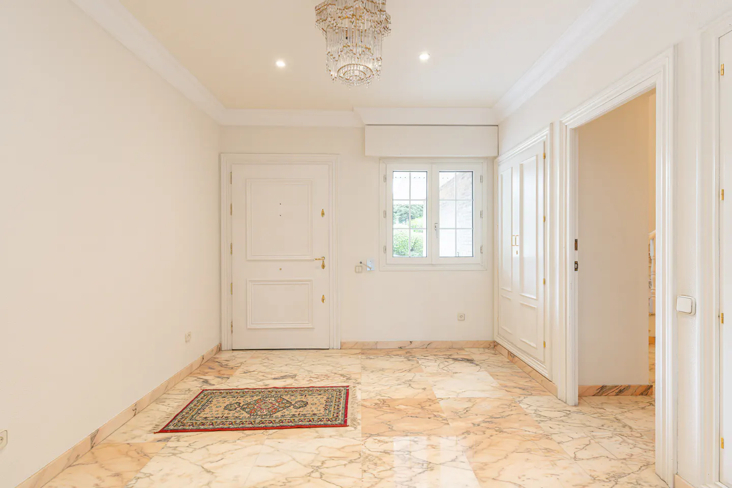 Bright foyer with marble floors, white walls, and a crystal chandelier. A rug sits near the front door, and a window lets in natural light.
