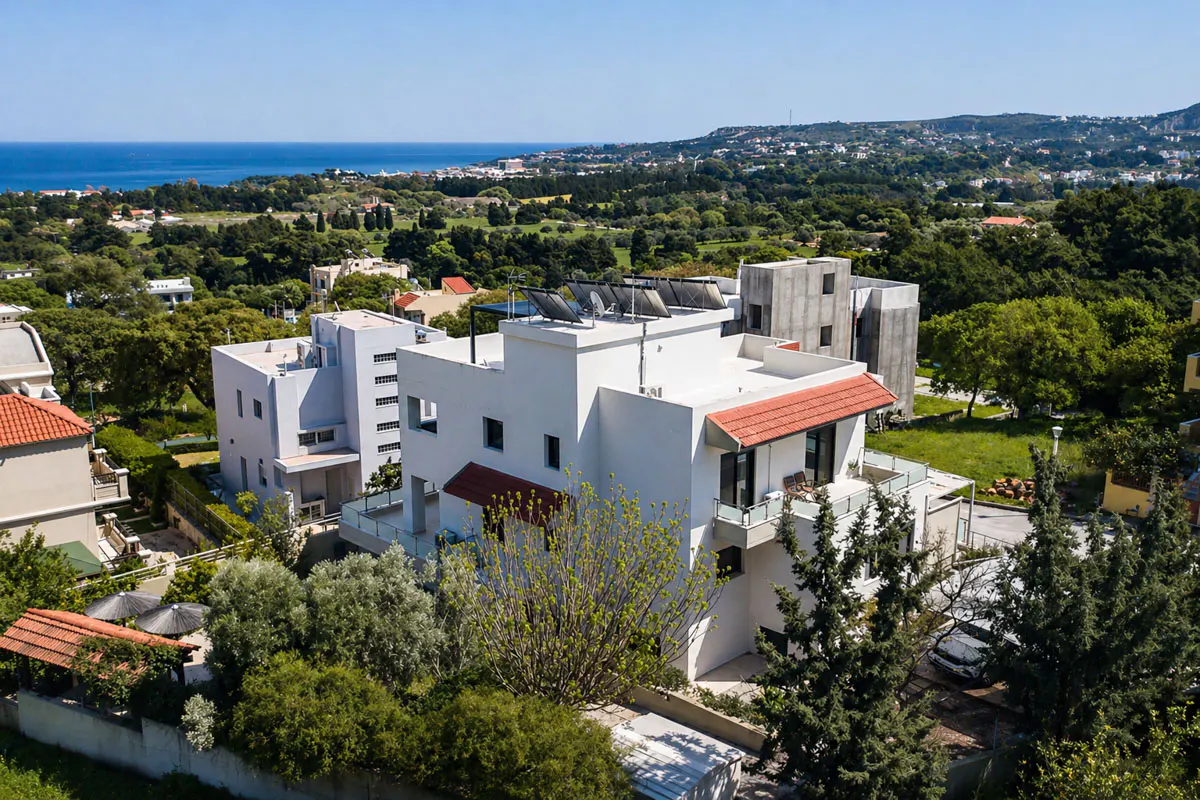 Aerial view of a modern white house with red roof accents, solar panels, and lush green surroundings near the ocean.