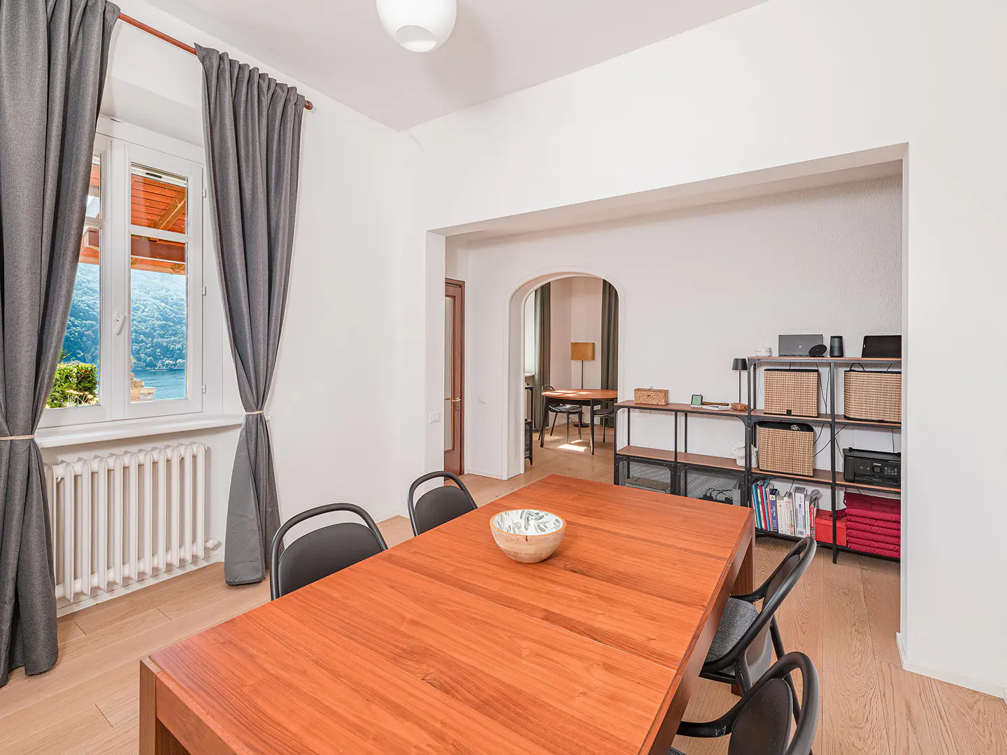 Bright dining room with a long wooden table, black chairs, and a window view of a lake. Shelves and an arched doorway lead to another room.