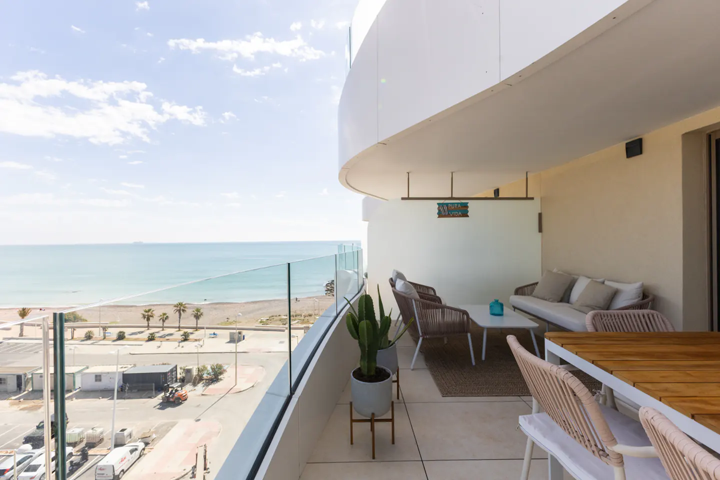 Balcony view with outdoor furniture, glass railing, and ocean backdrop. Cactus in a pot adds a touch of greenery. Sunny day.