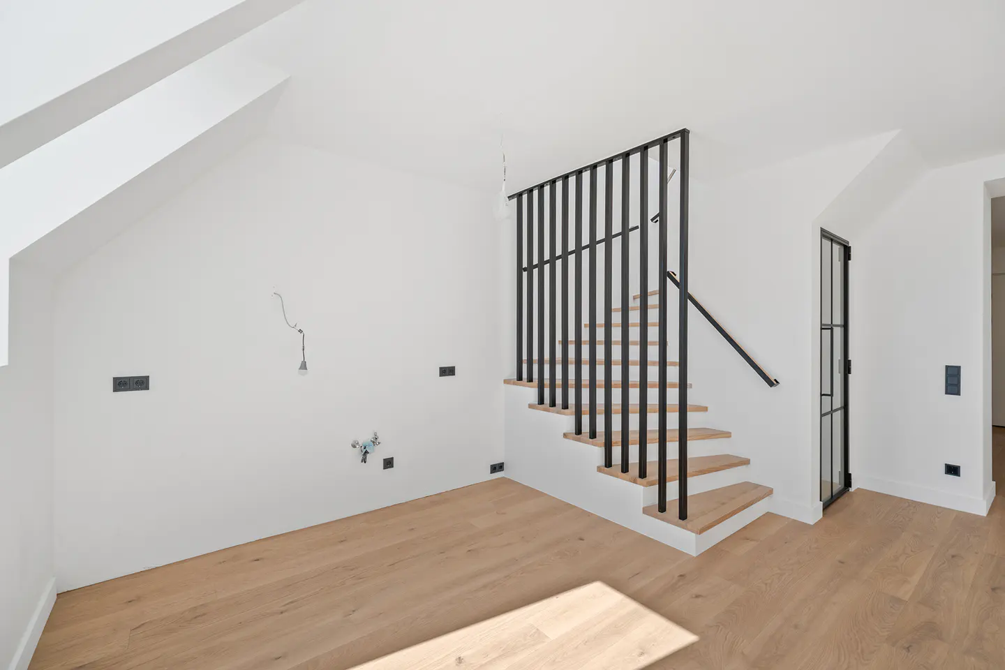 Bright room with light wood floors, white walls, and a black metal staircase. A black framed glass door is visible.