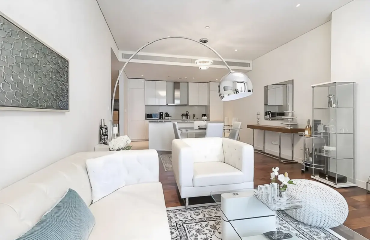 Bright, modern living room with white sofa, chair, and open kitchen. A silver arc lamp hangs over the seating area. Artwork adorns the wall.