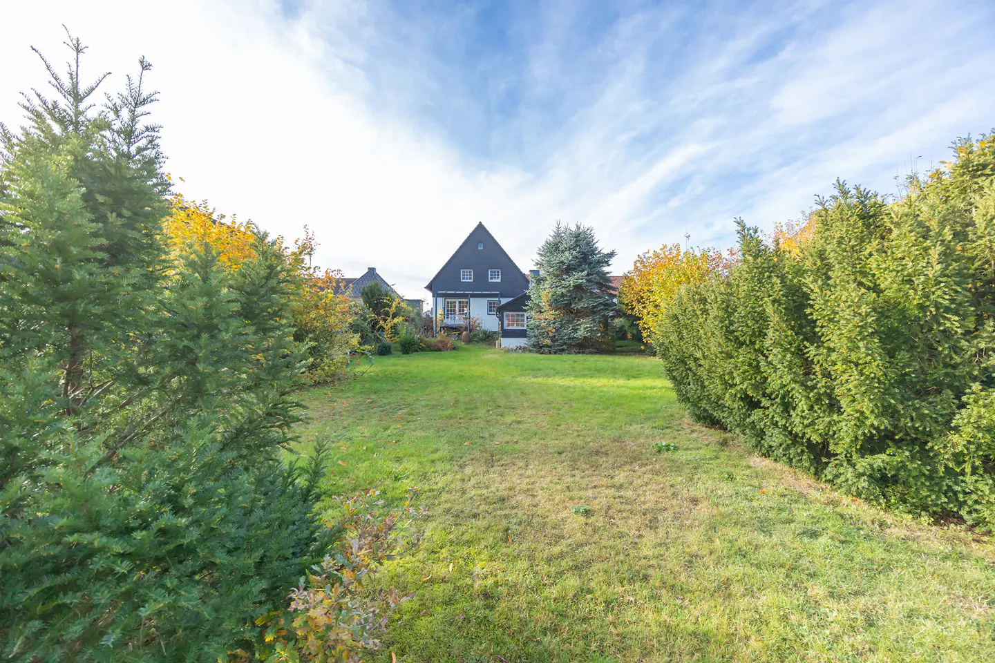 View of a house with a dark roof and white siding, framed by green trees and a large lawn under a blue sky.