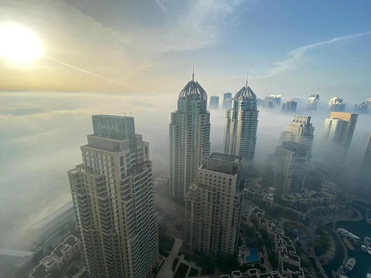 Aerial view of Dubai skyscrapers rising above a thick layer of morning fog under a bright, hazy sun.