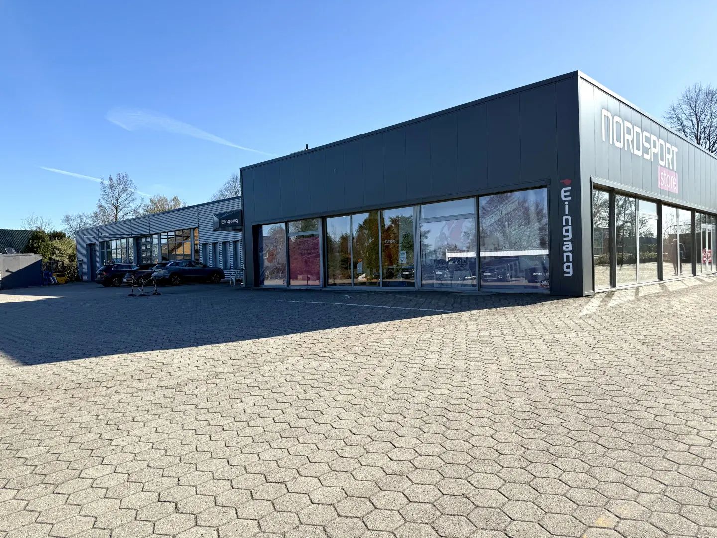Exterior view of Nordsport Store, a gray building with large windows, and "Eingang" (Entrance) sign. Cars parked in front.