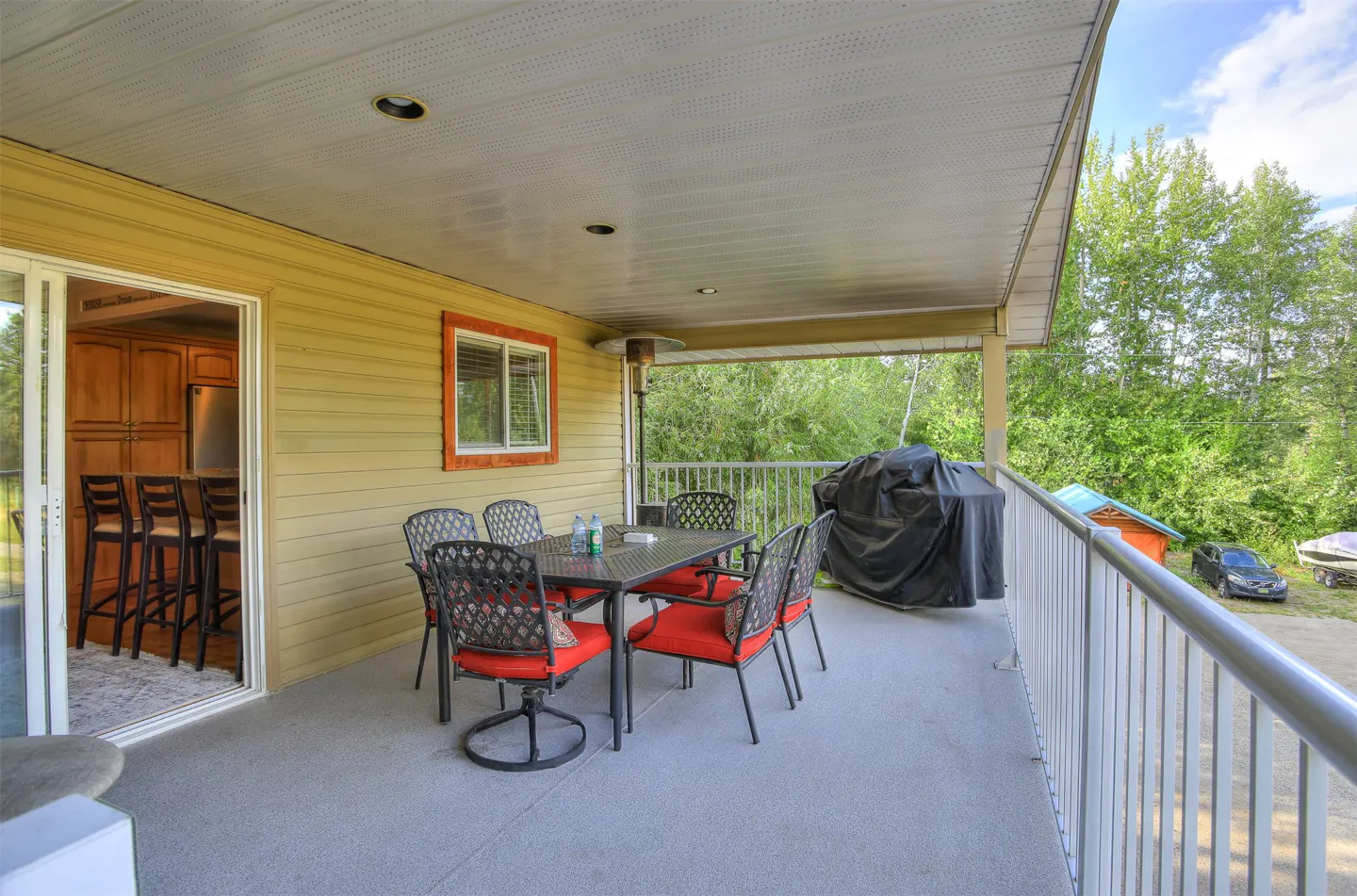Covered patio with table, chairs with red cushions, and grill. Sliding glass door leads to kitchen with bar stools. View of trees and sky.