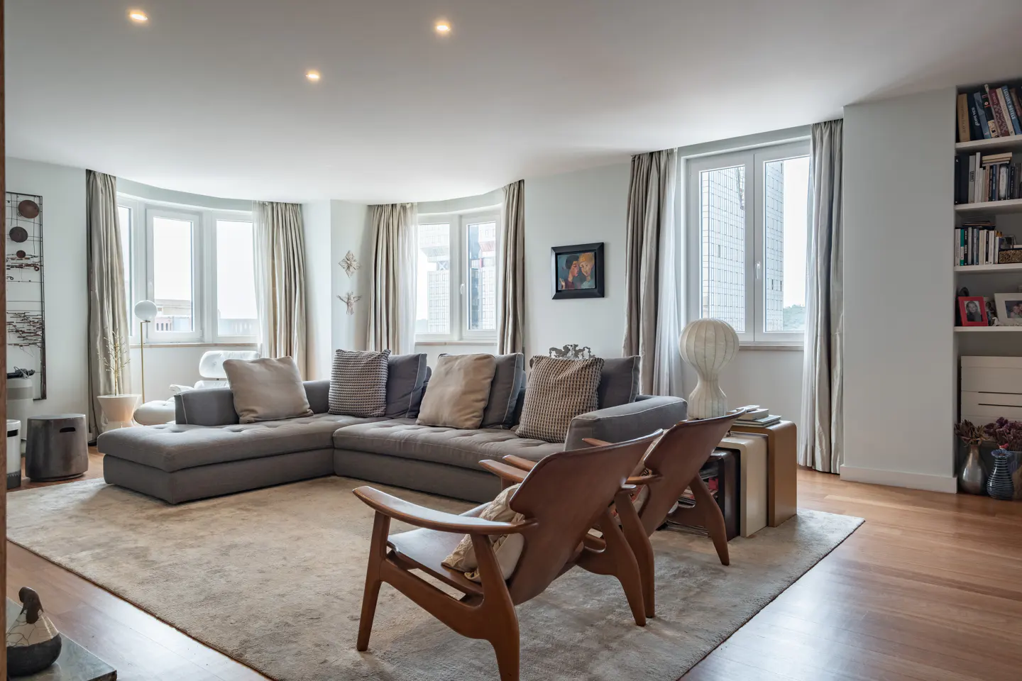 Bright living room with a gray sectional sofa, wood chairs, and large windows with curtains. A bookshelf is on the right.
