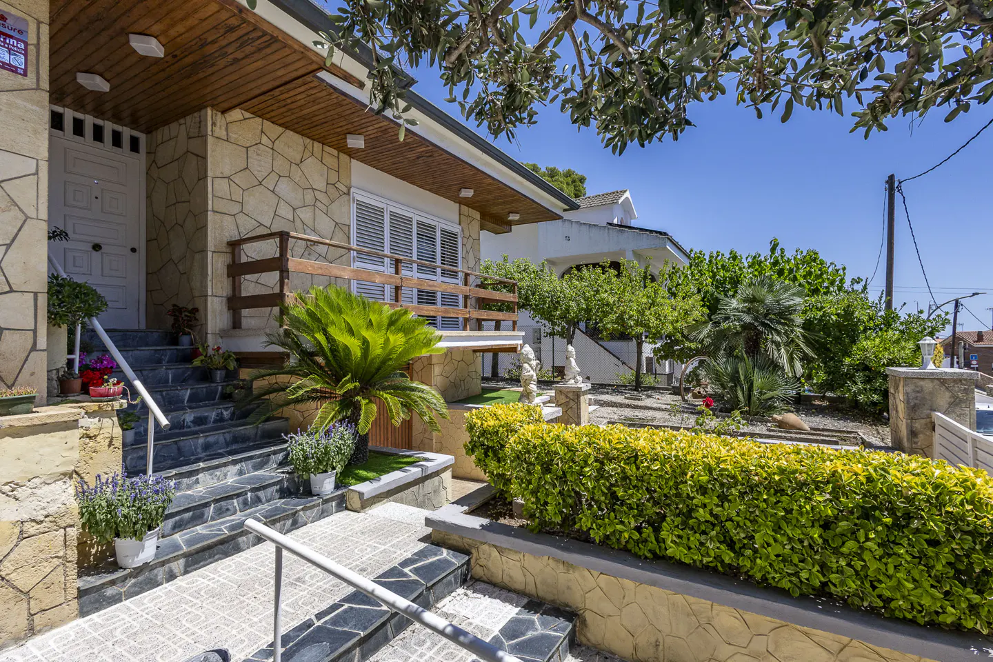 Stone house entrance with stairs, wooden balcony, lush green garden, and bright blue sky on a sunny day.