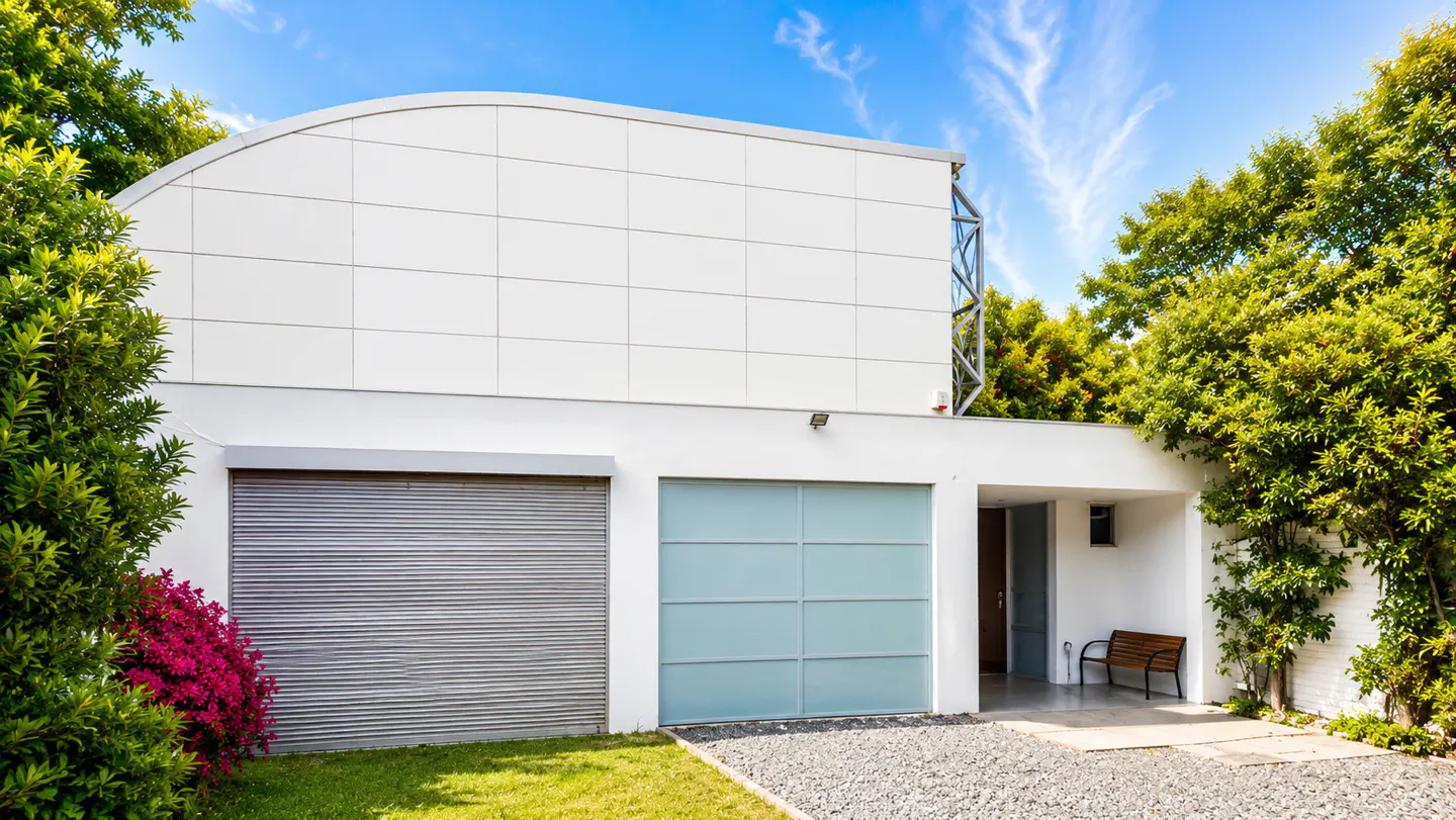 Modern white house with two garages, one gray and one blue, surrounded by green trees and a blue sky.